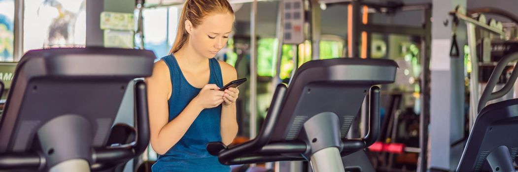 Young woman using phone while training at the gym. Woman sitting on exercising machine holding mobile phone. BANNER, LONG FORMAT