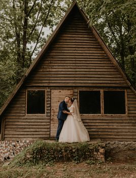 European Caucasian young man groom in blue suit and black-haired woman bride in white wedding dress with long veil and tiara on head. Newlyweds hugging standing near wooden small triangular house
