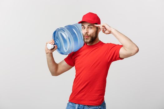Delivery Concept: Portrait of smiling bottled water delivery courier in red t-shirt and cap carrying tank of fresh drink isolated over grey background.