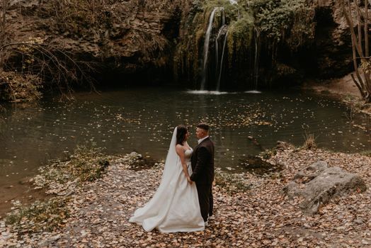 skinny bride in white wedding dress and long veil hugs groom in a suit. small pond lake river. summer-autumn sunset fallen orange leaves mist. waterfall Burbun in village Lysets Dunaivtsi Khmelnytskyi