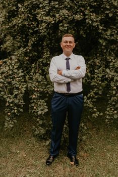 young blond man European curly-haired successful businessman groom in classic clothes white shirt and tie stands on street with his hands clasped together in nature on background of a green leafy wall