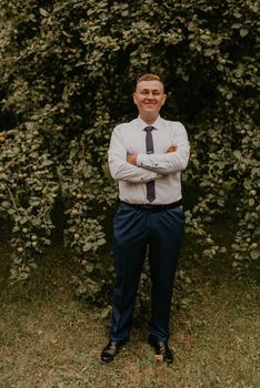 young blond man European curly-haired successful businessman groom in classic clothes white shirt and tie stands on street with his hands clasped together in nature on background of a green leafy wall