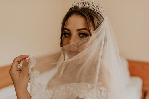 European Caucasian young black-haired woman bride in white wedding dress with long veil and tiara on head. girl bedroom in beige colors morning gatherings of preparing bride for holiday party wedding