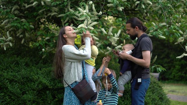 Happy family mother Father four Baby little siblings kids have fun sniffing flowers on trees enjoying summer holidays in garden park. Smiling parents children spending leisure time together sunset.