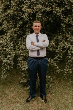 young blond man European curly-haired successful businessman groom in classic clothes white shirt and tie stands on street with his hands clasped together in nature on background of a green leafy wall