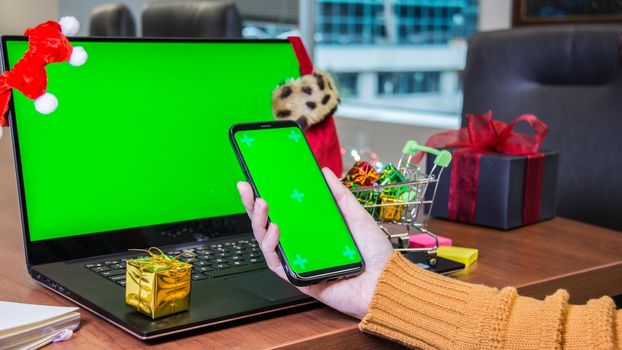 Closeup woman hand holding smartphone with green screen next to laptop with empty green screen and Christmas decoration, gift box on background. Holiday concept. Online shopping. Office. Mock-up.