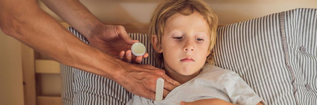 BANNER, LONG FORMAT Sick boy with thermometer laying in bed and father hand taking temperature. father checking temperature of her sick son who has thermometer. Sick child with fever and illness in bed.