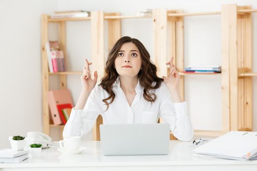 Portrait of happy young caucasian business woman sitting at the office desk and holding fingers crossed isolated over white background.