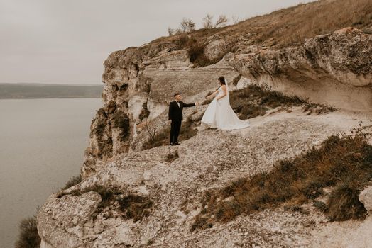 loving couple wedding newlyweds outdoor. bride in white dress long veil and groom in suit walk in summer fall on mountain above river. sunrise. man and woman on rocks above cliff. bakota ukraine