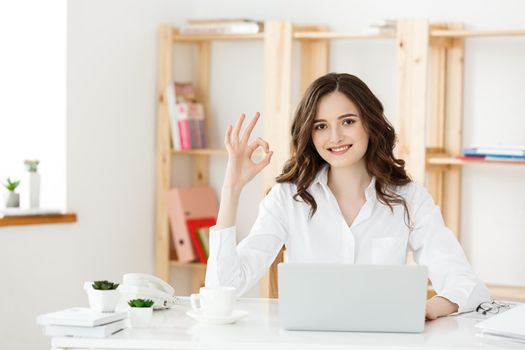 Young business woman showing ok sign sitting in the modern office with laptop