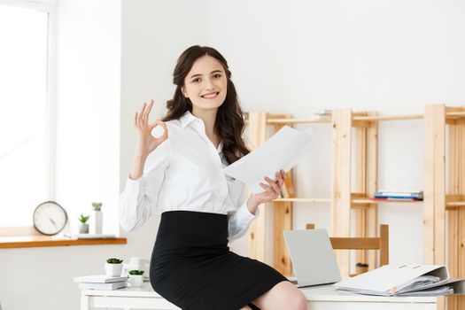 Businesswoman in formal wear holding documents and showing ok sign at modern office, copy space