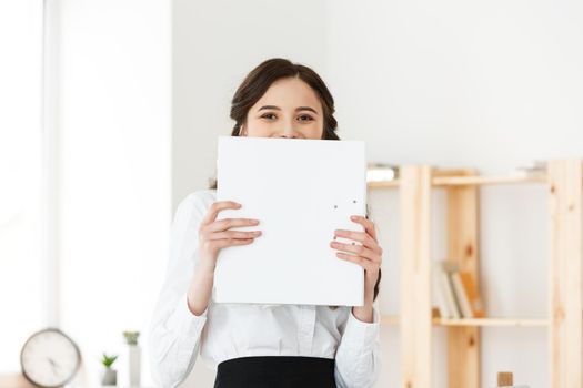 Young woman with surprised eyes peeking out from behind paper poster. Businesswoman holding big white banner in modern office