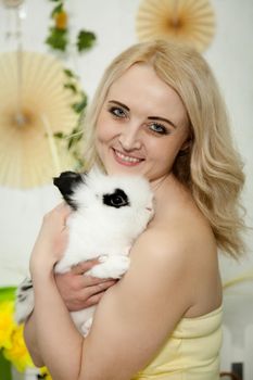 A lovely young blond woman standing in profile closeup and holding a young bunny in her arms. Beautiful blonde girl holding a white rabbit in her arms. Easter bunny sitting in the arms of a girl