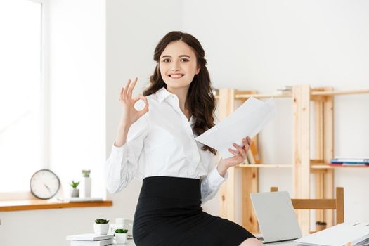 Businesswoman in formal wear holding documents and showing ok sign at modern office, copy space