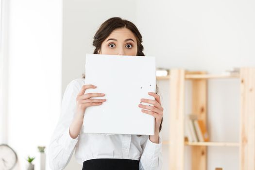 Young woman with surprised eyes peeking out from behind paper poster. Businesswoman holding big white banner in modern office