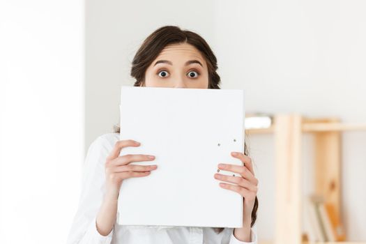 Young woman with surprised eyes peeking out from behind paper poster. Businesswoman holding big white banner in modern office