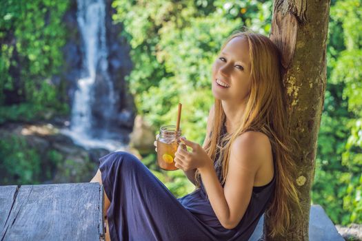 Closeup portrait image of a beautiful woman drinking ice tea with feeling happy in green nature and waterfall garden background.
