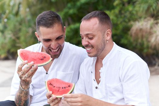 Portrait of a gay couple sitting on a beach while smiling and holing a piece of watermelon