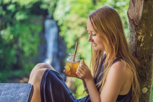 Closeup portrait image of a beautiful woman drinking ice tea with feeling happy in green nature and waterfall garden background.