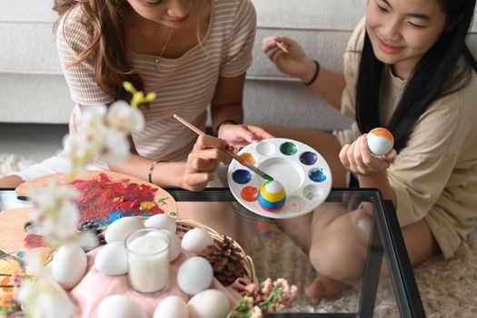 Little cute girl and mother are coloring eggs preparing for Easter celebration at home together.