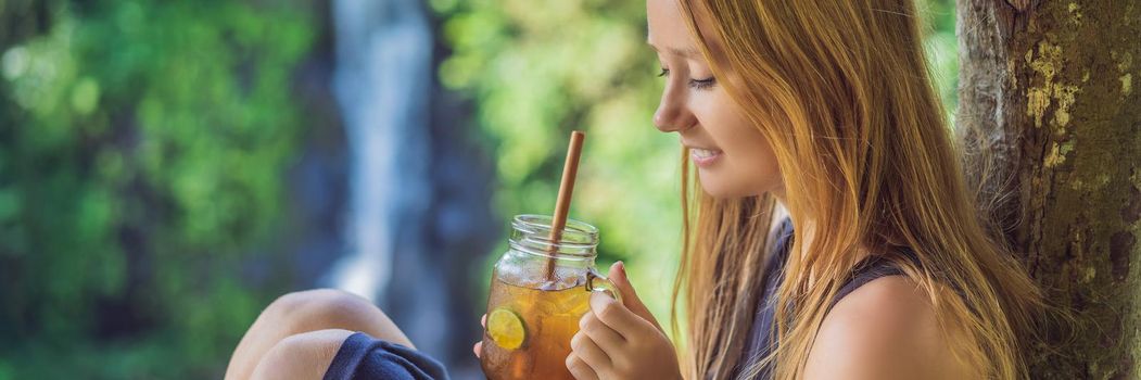 Closeup portrait image of a beautiful woman drinking ice tea with feeling happy in green nature and waterfall garden background. BANNER, LONG FORMAT