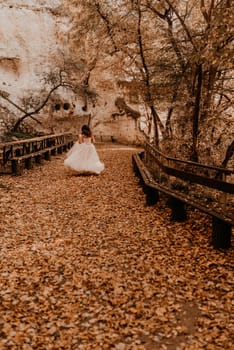 woman bride in white wedding dress with hairstyle makeup and crown on her head walks through autumn forest on fallen orange leaves