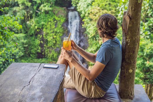 Closeup portrait image of a beautiful man drinking ice tea with feeling happy in green nature and waterfall garden background.