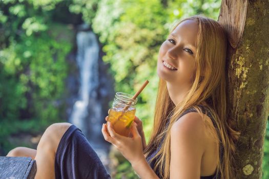 Closeup portrait image of a beautiful woman drinking ice tea with feeling happy in green nature and waterfall garden background.
