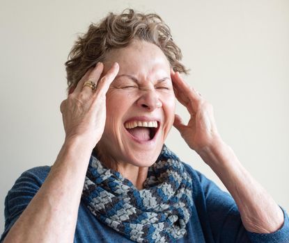 Head and shoulders view of older woman in blue clothing with hands on head screaming (selective focus)