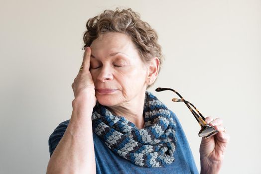 Head and shoulders view of older woman in blue clothing rubbing eyes and holding glasses (selective focus)