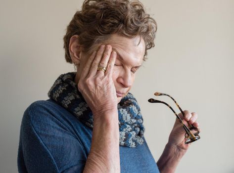 Head and shoulders view of older woman in blue clothing rubbing eyes and holding glasses (selective focus)