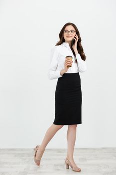 Full length Portrait of beautiful young business woman in formal wear walking and talking for cell phone with takeaway coffee in hand isolated over gray background.