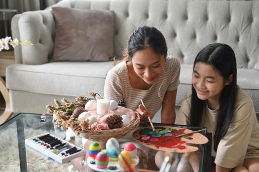 Beautiful young mother helping her daughter painting Easter eggs. Easter, holidays and people concept.