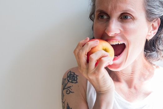 Close up of natural looking middle aged woman with grey hair biting into red apple (selective focus)