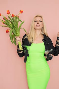 Young elegant woman in trendy jeans jacket. Blonde hair, green dress, isolated over pink background, studio shot. Fashion spring lookbook. Model woman with red tulips in her hands