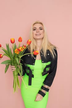 Young elegant woman in trendy jeans jacket. Blonde hair, green dress, isolated over pink background, studio shot. Fashion spring lookbook. Model woman with red tulips in her hands