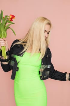 Young elegant woman in trendy jeans jacket. Blonde hair, green dress, isolated over pink background, studio shot. Fashion spring lookbook. Model woman with red tulips in her hands