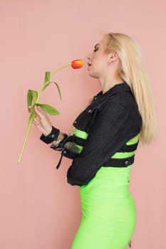 Young elegant woman in trendy jeans jacket. Blonde hair, green dress, isolated over pink background, studio shot. Fashion spring lookbook. Model woman with red tulips in her hands