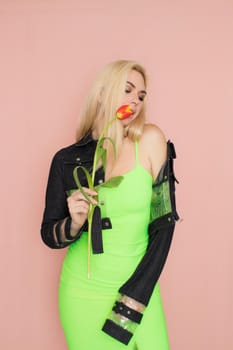 Young elegant woman in trendy jeans jacket. Blonde hair, green dress, isolated over pink background, studio shot. Fashion spring lookbook. Model woman with red tulips in her hands