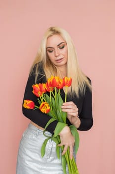 Fashion model woman in fashionable clothes on pink background. Wearing stylish clothing, black blouse, silver skirt. Posing in studio. Holding red tulips in her hands