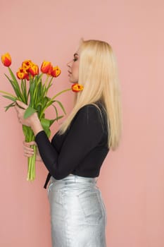 Fashion model woman in fashionable clothes on pink background. Wearing stylish clothing, black blouse, silver skirt. Posing in studio. Holding red tulips in her hands