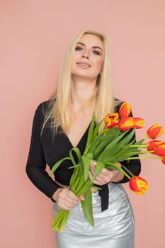 Fashion model woman in fashionable clothes on pink background. Wearing stylish clothing, black blouse, silver skirt. Posing in studio. Holding red tulips in her hands