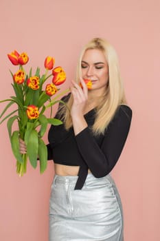 Fashion model woman in fashionable clothes on pink background. Wearing stylish clothing, black blouse, silver skirt. Posing in studio. Holding red tulips in her hands