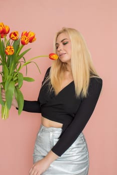 Fashion model woman in fashionable clothes on pink background. Wearing stylish clothing, black blouse, silver skirt. Posing in studio. Holding red tulips in her hands