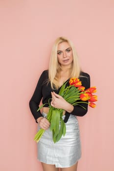 Fashion model woman in fashionable clothes on pink background. Wearing stylish clothing, black blouse, silver skirt. Posing in studio. Holding red tulips in her hands