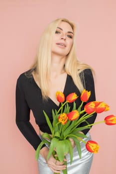 Fashion model woman in fashionable clothes on pink background. Wearing stylish clothing, black blouse, silver skirt. Posing in studio. Holding red tulips in her hands