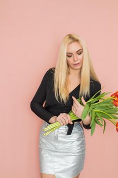 Fashion model woman in fashionable clothes on pink background. Wearing stylish clothing, black blouse, silver skirt. Posing in studio. Holding red tulips in her hands