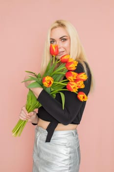 Fashion model woman in fashionable clothes on pink background. Wearing stylish clothing, black blouse, silver skirt. Posing in studio. Holding red tulips in her hands