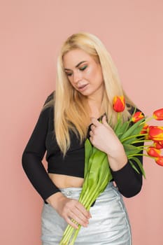 Fashion model woman in fashionable clothes on pink background. Wearing stylish clothing, black blouse, silver skirt. Posing in studio. Holding red tulips in her hands