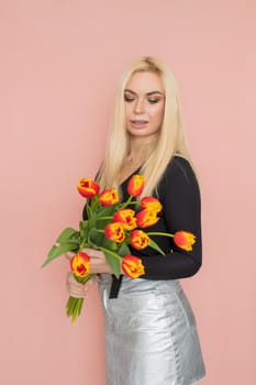 Fashion model woman in fashionable clothes on pink background. Wearing stylish clothing, black blouse, silver skirt. Posing in studio. Holding red tulips in her hands